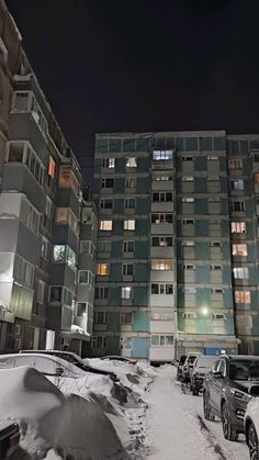 several cars parked in front of an apartment building at night with snow on the ground