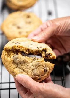 a person holding a chocolate chip cookie in front of some cookies on a cooling rack