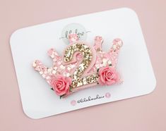 a pink and gold crown brooch with two roses on the front, sitting on top of a white card