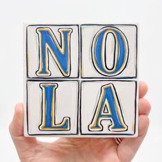 a hand holding up a ceramic block with the word nola on it in blue and yellow letters