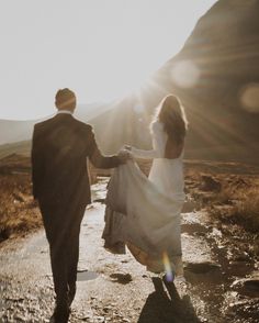 a bride and groom holding hands walking down a path in the mountains with sun shining on them