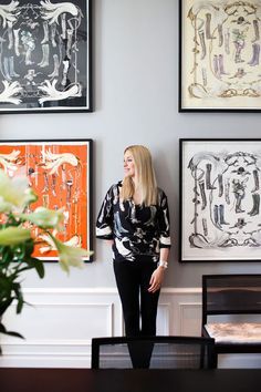 a woman standing in front of some paintings on the wall next to a vase with flowers