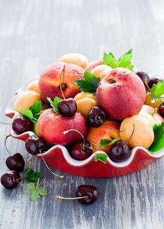 a red bowl filled with lots of fruit on top of a wooden table next to cherries