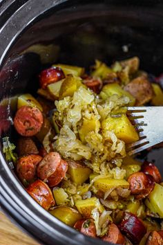 a fork in a crock pot filled with cooked sausage, potatoes and peppers on a wooden table