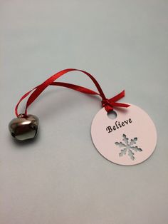 a bell ornament hanging from a red ribbon on a white surface with the word believe written in it