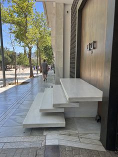 two white benches sitting on the side of a building next to a sidewalk with people walking by