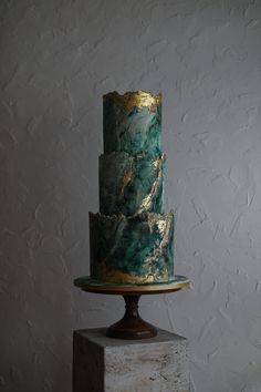 a three tiered cake with green and gold icing on a marble pedestal in front of a white wall