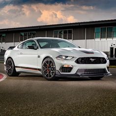 a white mustang parked in front of a building on a race track with clouds overhead