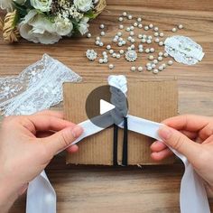 two hands holding scissors over a cardboard box with white ribbon and flowers in the background