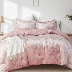 a bed with pink comforter and pillows in a room next to a plant on the floor