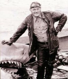 a man standing next to a shark on top of a boat with it's mouth open