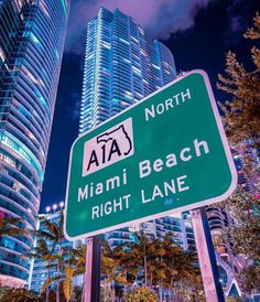 a green street sign sitting in front of tall buildings at night with the words miami beach right lane written on it