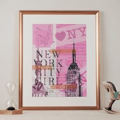 a framed art print with the words new york city in pink and black, on a shelf next to an hourglass