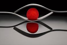 a red ball sitting on top of a metal fork next to a shiny surface with reflections