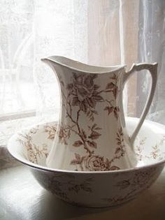 a white and brown flowered pitcher sitting on top of a saucer in front of a window