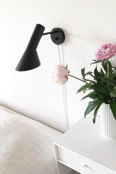 a white vase with pink flowers sitting on a table next to a black wall light