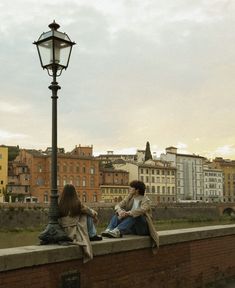 two women sitting on a brick wall next to a street light and lamp post with buildings in the background