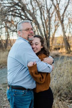 an older man and young woman hugging each other in front of some trees with no leaves