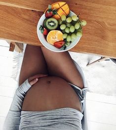 a pregnant woman sitting at a table with fruit in a bowl on her stomach, looking down