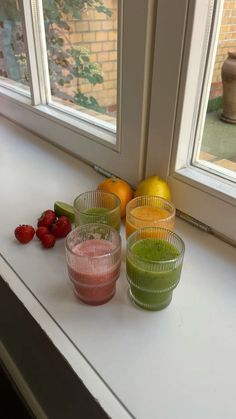 four glasses filled with different colored drinks sitting on a window sill next to fruit