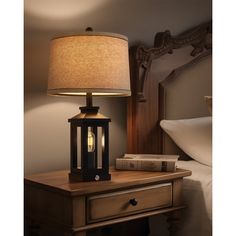 a lamp on a night stand next to a nightstand with a book and a bed in the background