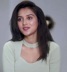 a woman with long black hair wearing a white shirt and looking at the camera while standing in front of a gray wall