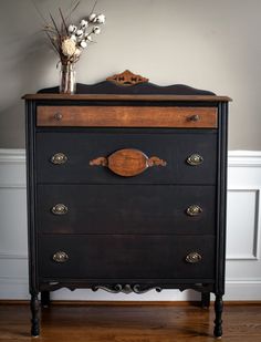 a black dresser with wooden drawers and flowers on top, in front of a white wall