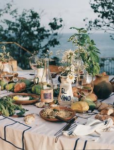 a table topped with plates and glasses filled with food next to the ocean on a sunny day