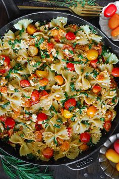 a skillet filled with pasta and vegetables on top of a wooden table next to utensils