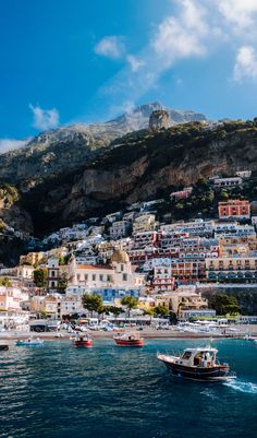 boats are on the water in front of some buildings and cliffs, with mountains in the background