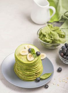 a stack of green pancakes with bananas and blueberries on the plate next to some leaves