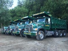 a row of green dump trucks parked next to each other on a dirt road with trees in the background