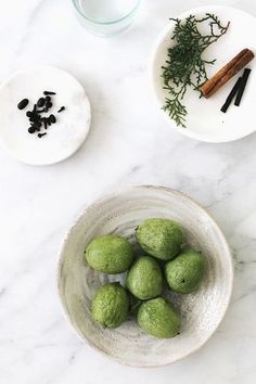 some green balls in a white bowl on a marble table with cinnamon sticks and spices