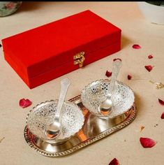 two silver serving dishes with spoons on a tray next to a red box and rose petals