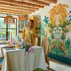 a woman standing next to a table in front of a wall with flowers on it