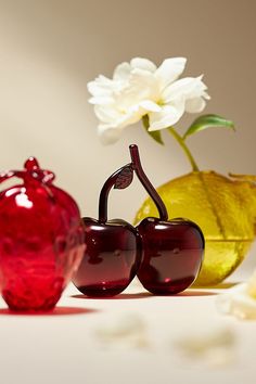 two red glass cherries sitting next to a yellow vase with white flowers in it