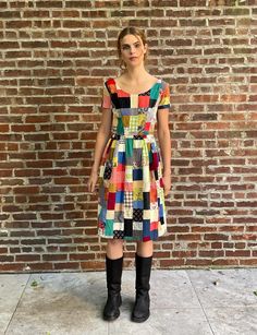 a woman standing in front of a brick wall wearing a multicolored dress and black boots