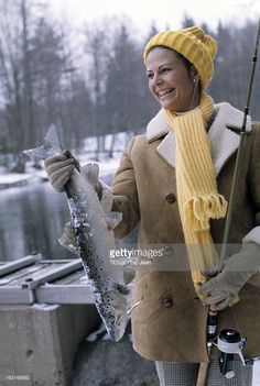 a woman holding a fish while wearing a yellow hat and scarf on the side of a lake