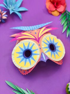 an owl mask made out of paper on a purple surface with flowers and leaves around it