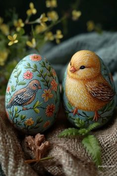 two painted eggs sitting next to each other on top of a cloth covered tablecloth