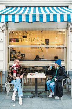 two people sitting at a table in front of a store with blue and white awnings