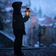 a person holding a lantern in the dark with snow falling on them and buildings in the background