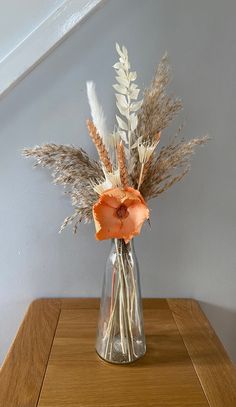 an orange flower in a glass vase on a wooden table next to some dry grass