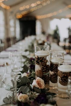 pine cones and candles are sitting on a long table with white linens, purple flowers and greenery