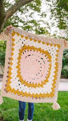 a woman holding up a crocheted blanket in front of a tree on the grass