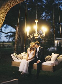 a bride and groom are sitting on a swing in the grass under a chandelier