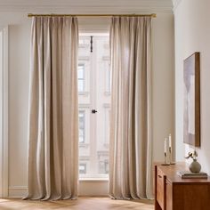 a living room with white walls and wooden flooring next to a large window covered in beige drapes