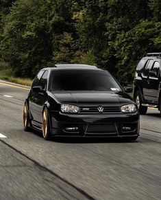 two black cars driving down the road next to each other in front of some trees