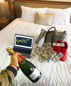 a person holding a bottle of champagne next to money and a laptop on a bed