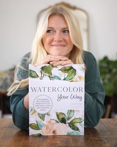 a woman sitting at a table holding up a box with watercolor your way written on it